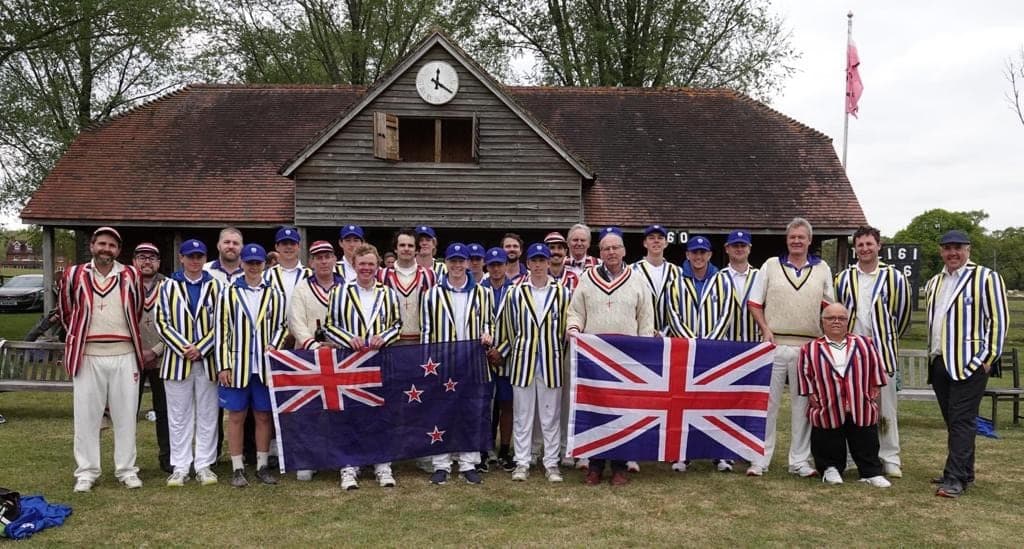 LNZCC and opposition players in club blazers with the New Zealand and Union flags in front of a wooden pavilion